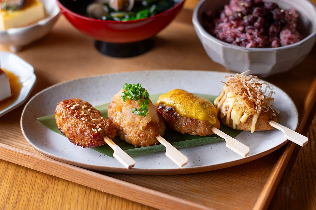 Four assorted grilled skewered bites on a plate with rice and side dishes