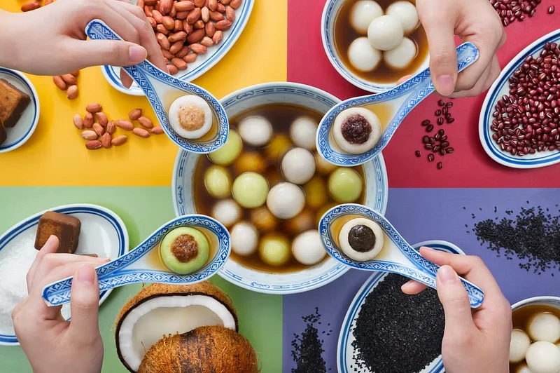 Hands holding decorated spoons over a bowl of colorful round sweets and assorted ingredients