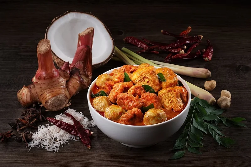 Bowl of orange curry with dumplings surrounded by chilies, coconut, lemongrass, and herbs